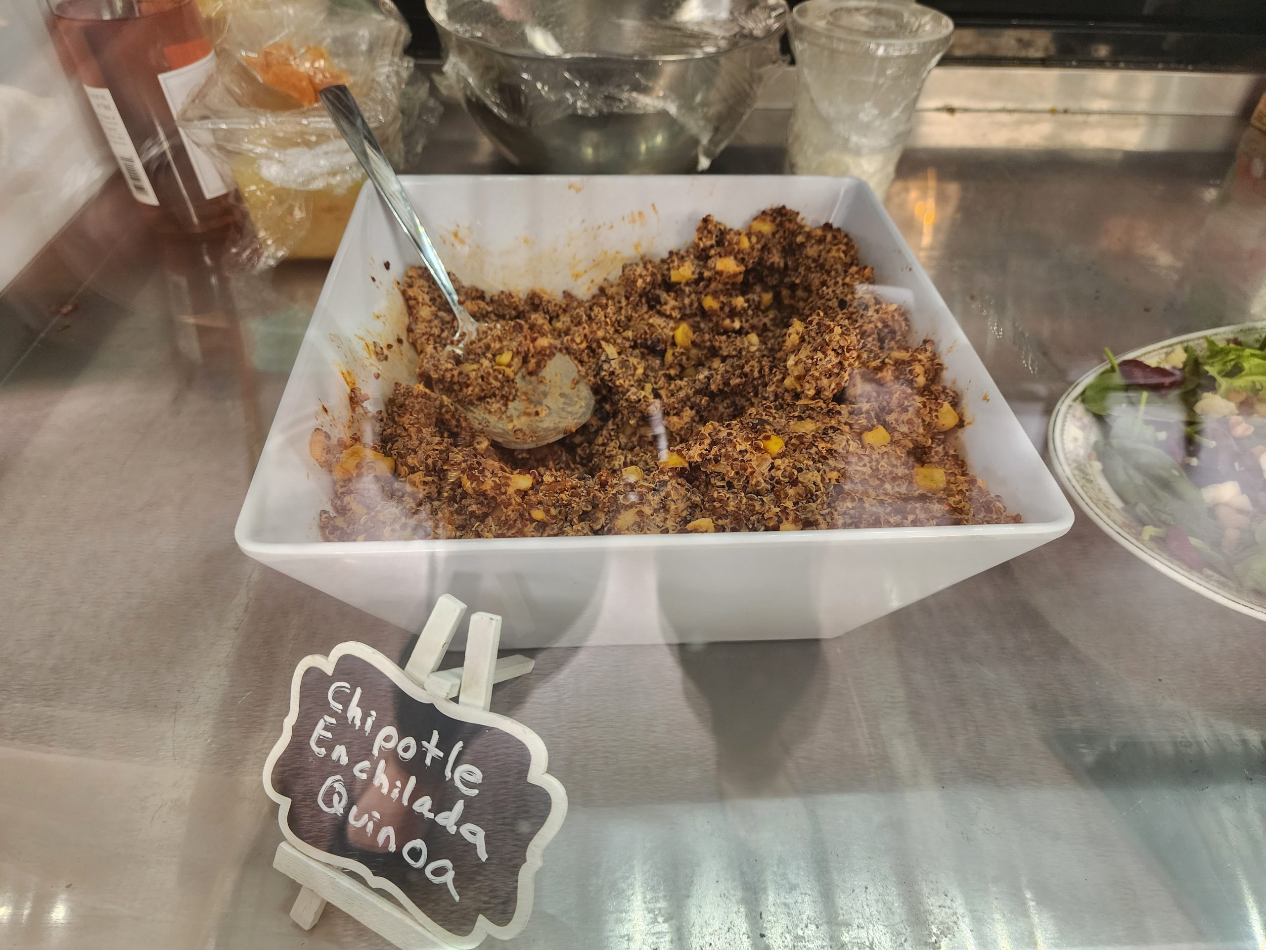 Quinoa salad in a white tray with a serving spoon on a stainless-steel counter.