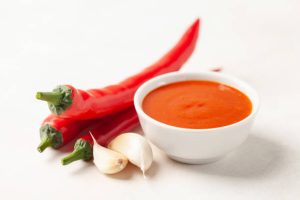 Bowl of red sriracha sauce with red chili peppers and garlic cloves.