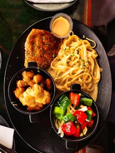 A delectable gourmet meal featuring pasta, breaded cutlet, and fresh vegetables served on a black plate.