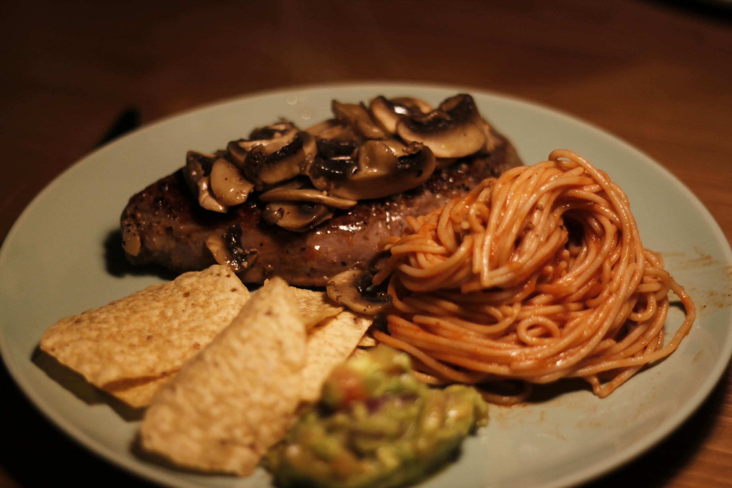 A gourmet beef dinner featuring steak with mushrooms, spaghetti, chips, and guacamole.