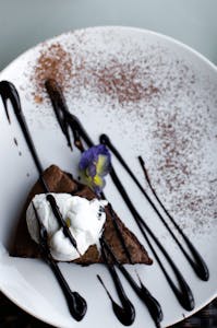 A gourmet chocolate dessert slice with whipped cream and edible flower on a ceramic plate, perfect for foodies.