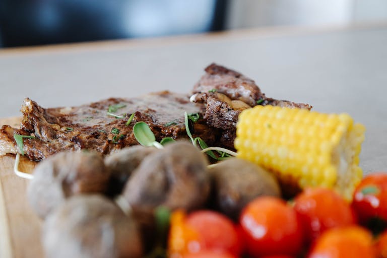 Close-up of a juicy grilled steak with cherry tomatoes, potatoes, and corn on a wooden platter.