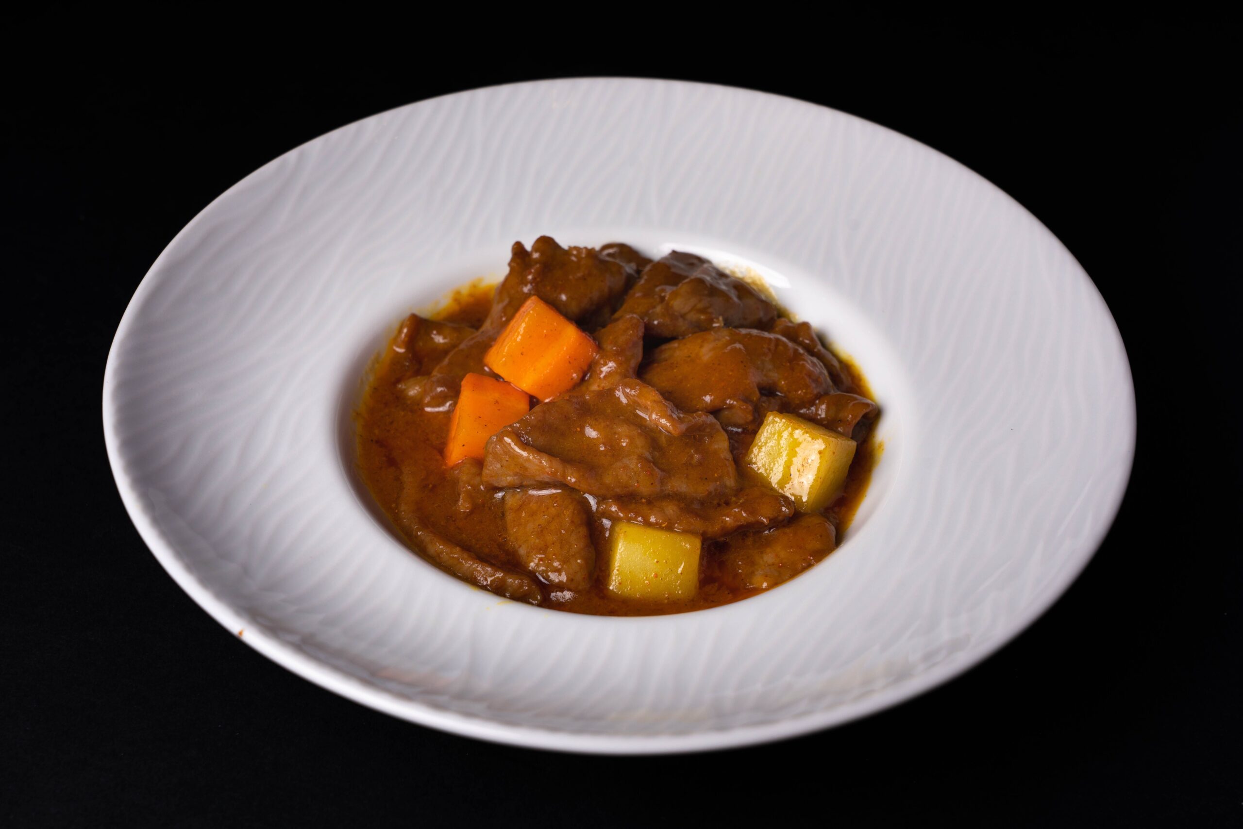 Beef pieces, orange carrot and pale potato chunks in a thick golden-brown sauce in a white bowl. Hidden spices, exact meat cut and the original recipe are not identifiable.