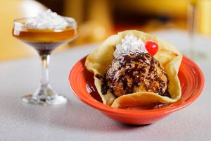 Fried ice cream in a tortilla shell topped with chocolate sauce, whipped cream, and a cherry, with a flan dessert in a glass beside it.