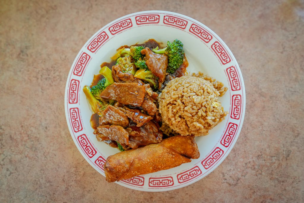 Delicious Chinese beef and broccoli with fried rice and egg roll on a decorative plate.