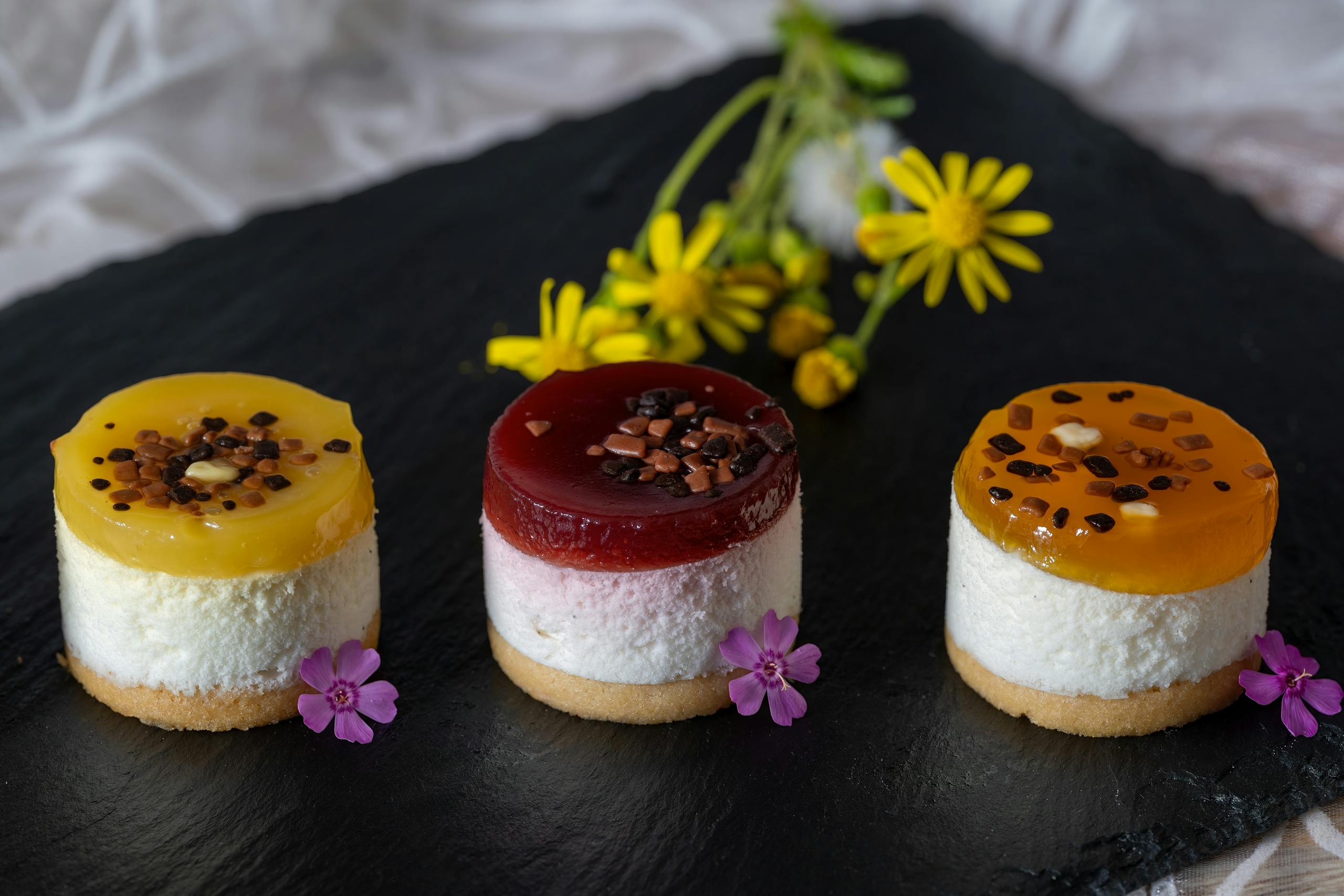 Delight in colorful mini fruit cheesecakes, beautifully garnished with flowers on a dark slate platter.