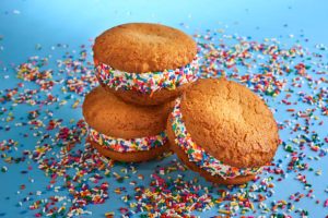 Three ice cream cookie sandwiches on a blue surface with colorful sprinkles.