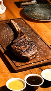 Juicy grilled tomahawk steak on a wooden board with three dipping sauces.