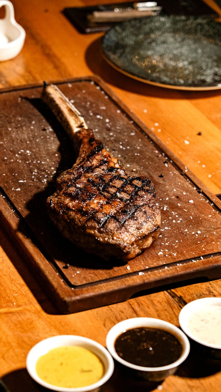 Juicy grilled tomahawk steak on a wooden board with three dipping sauces.