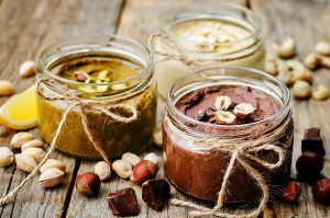 Glass jars of nut butter with pistachios, hazelnuts, and cashews on a wooden surface.
