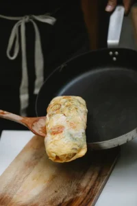 Rolled omelet lifted from a frying pan with a wooden spatula.