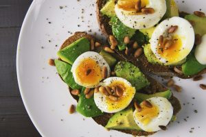 Avocado toast topped with soft-boiled eggs and pine nuts on a white plate.