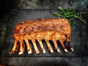 Herb-crusted rack of lamb on a black serving board with fresh herbs.