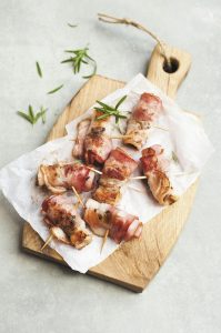 Bacon-wrapped chicken bites on parchment-lined wooden board with rosemary.