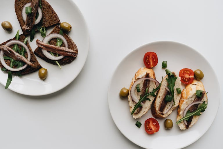 Tasty bruschetta topped with anchovies, arugula, and olives on white plates.