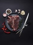Top view of raw T-Bone steak with spices, garlic, and cutlery on dark background.