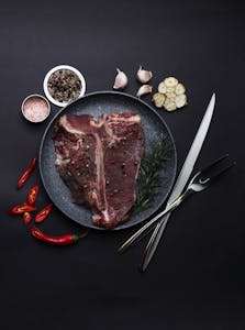 Top view of raw T-Bone steak with spices, garlic, and cutlery on dark background.