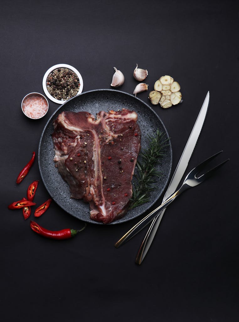 Top view of raw T-Bone steak with spices, garlic, and cutlery on dark background.