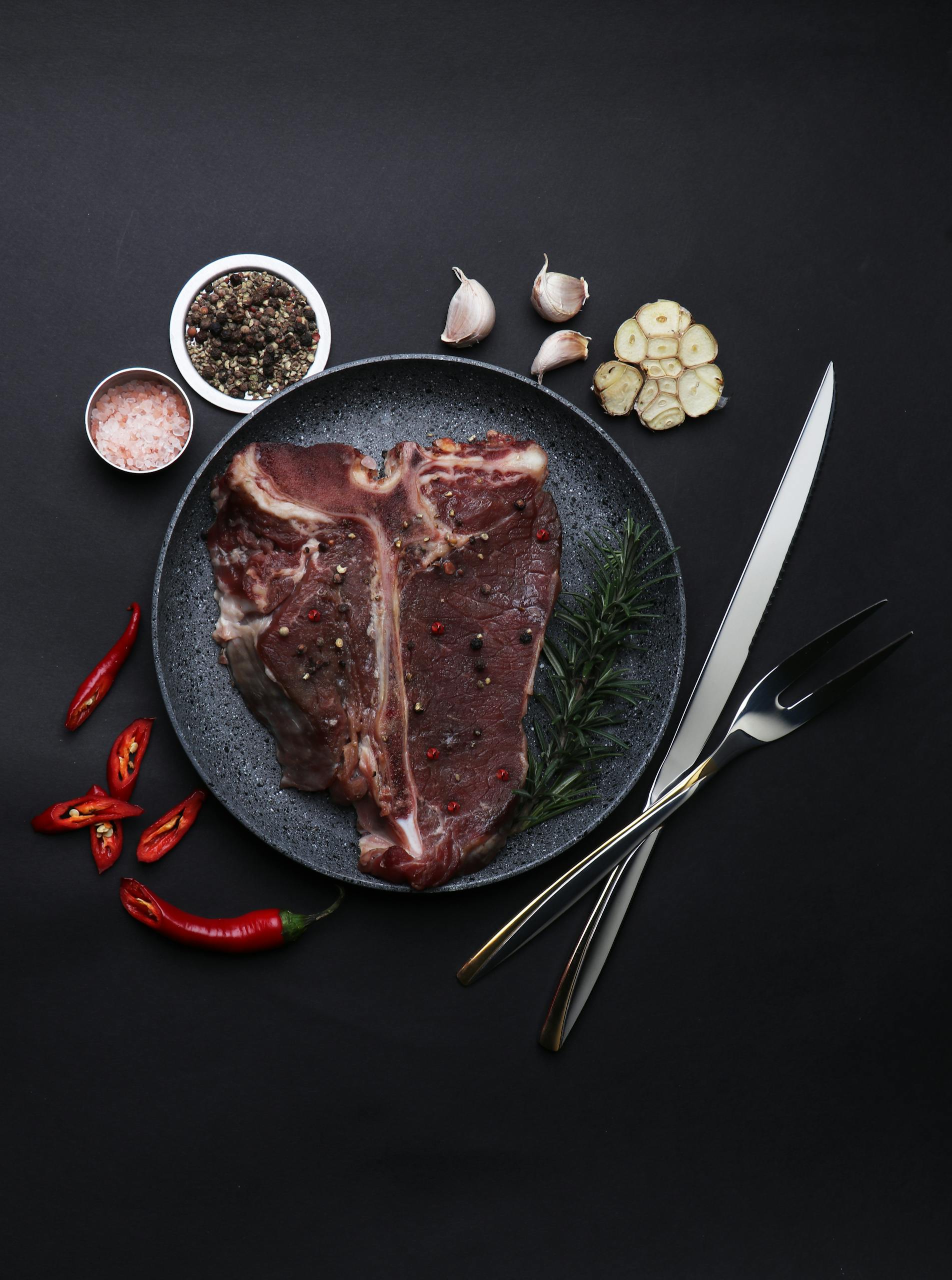 Top view of raw T-Bone steak with spices, garlic, and cutlery on dark background.