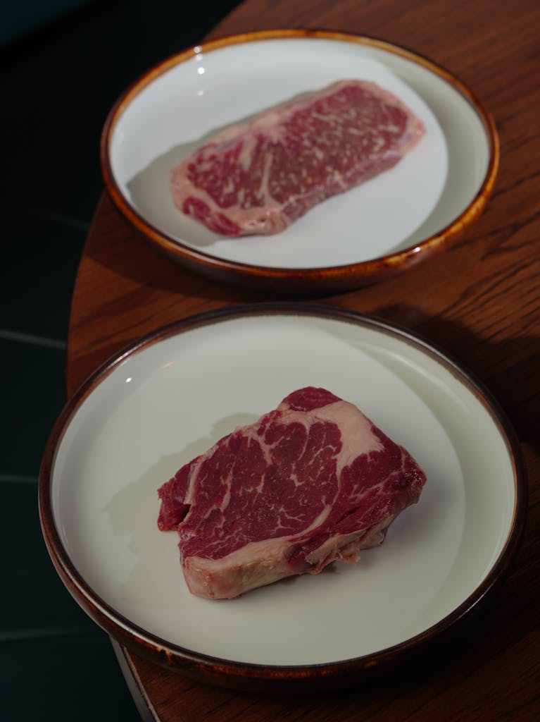 Two raw steak cuts on ceramic plates on a wooden table, ready for cooking.